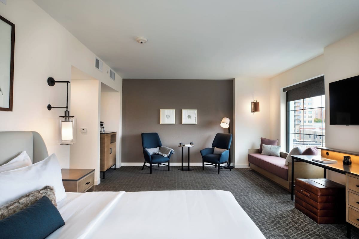 A hotel room with a large bed, two armchairs, a small table, a sofa, a desk, and a TV.