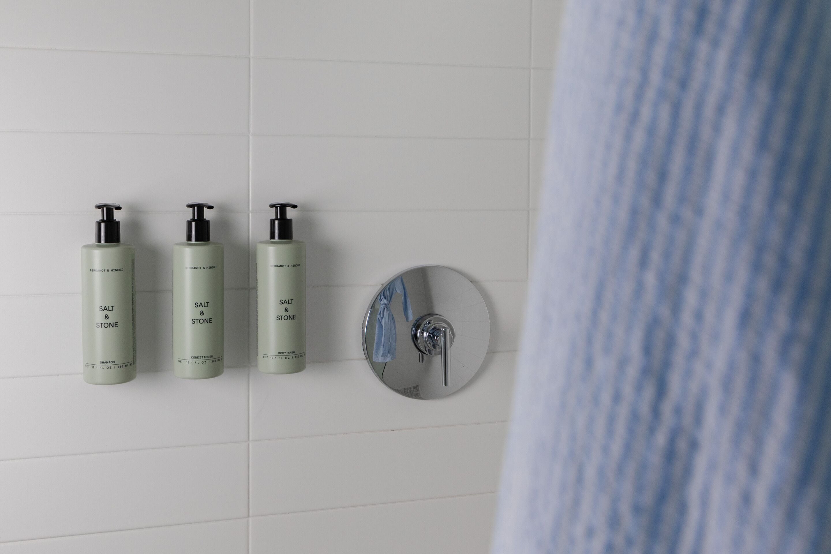 Three bottles of shampoo and conditioner on a white tiled wall.