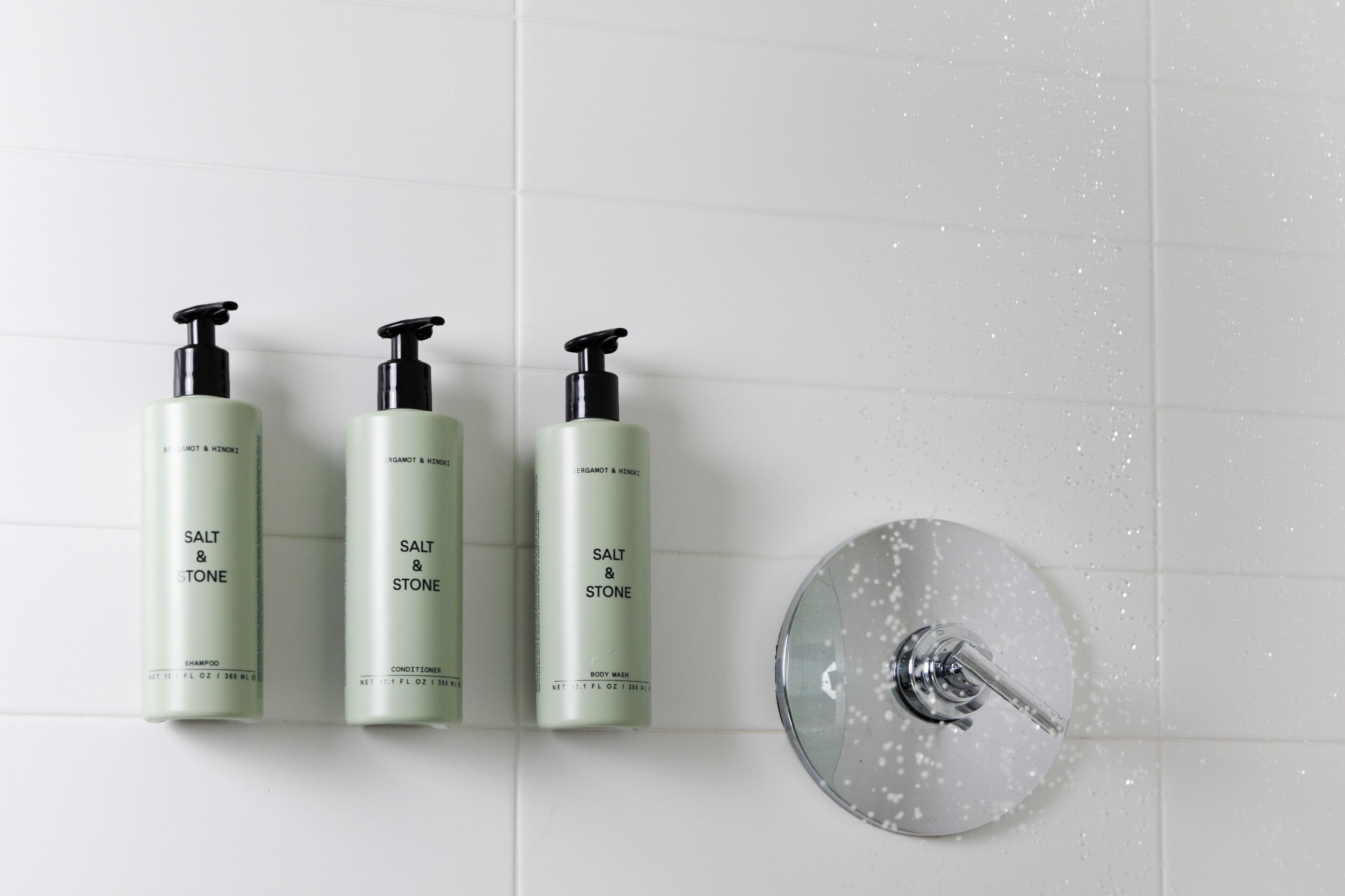 Trois bouteilles de shampoing, d’après-shampoing et de gel douche de la marque « Salt & Stone », posées sur un mur carrelé.