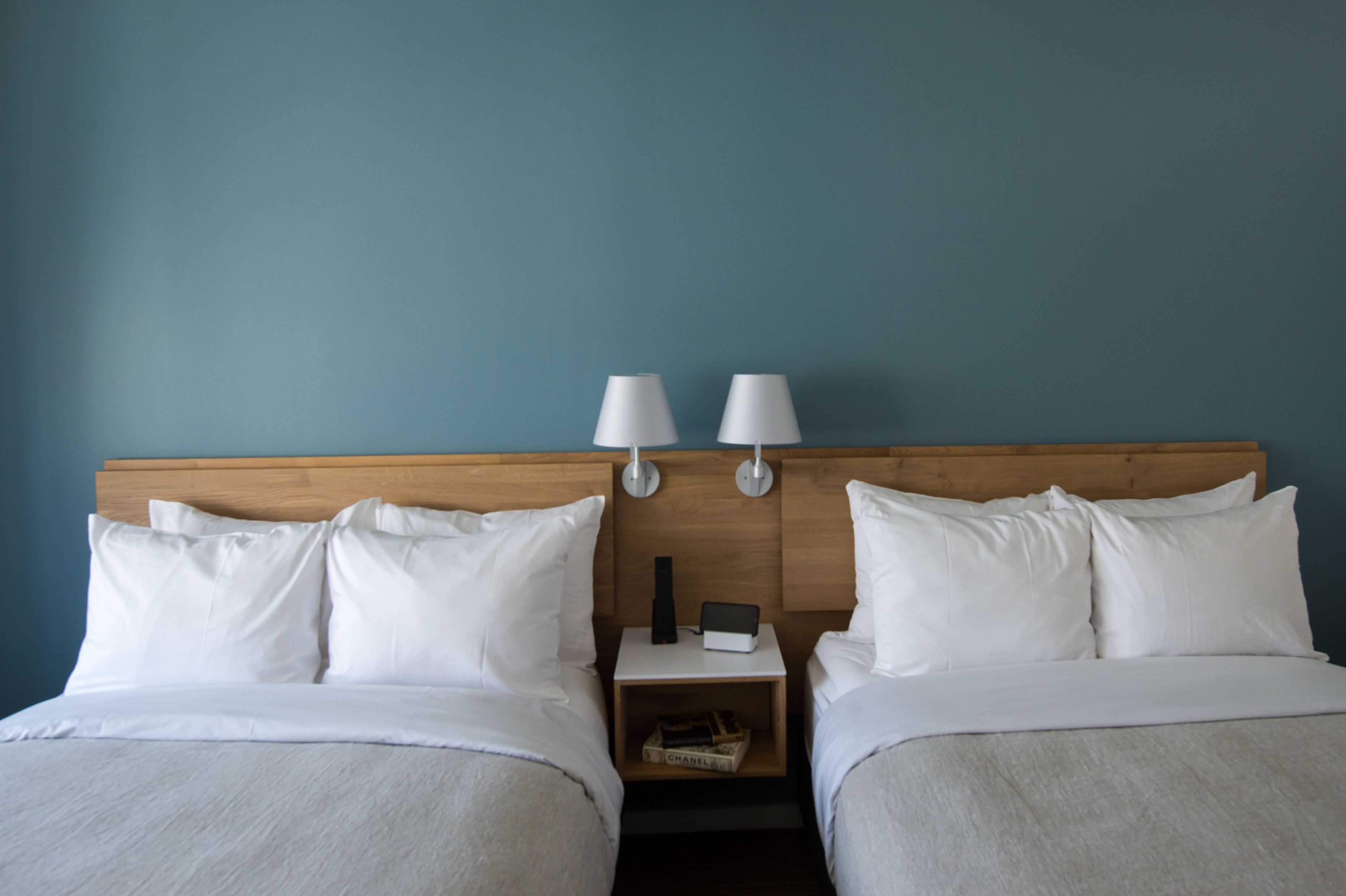 A hotel room with two beds, each with white bedding and pillows, a wooden headboard, and bedside tables with lamps and a small box.