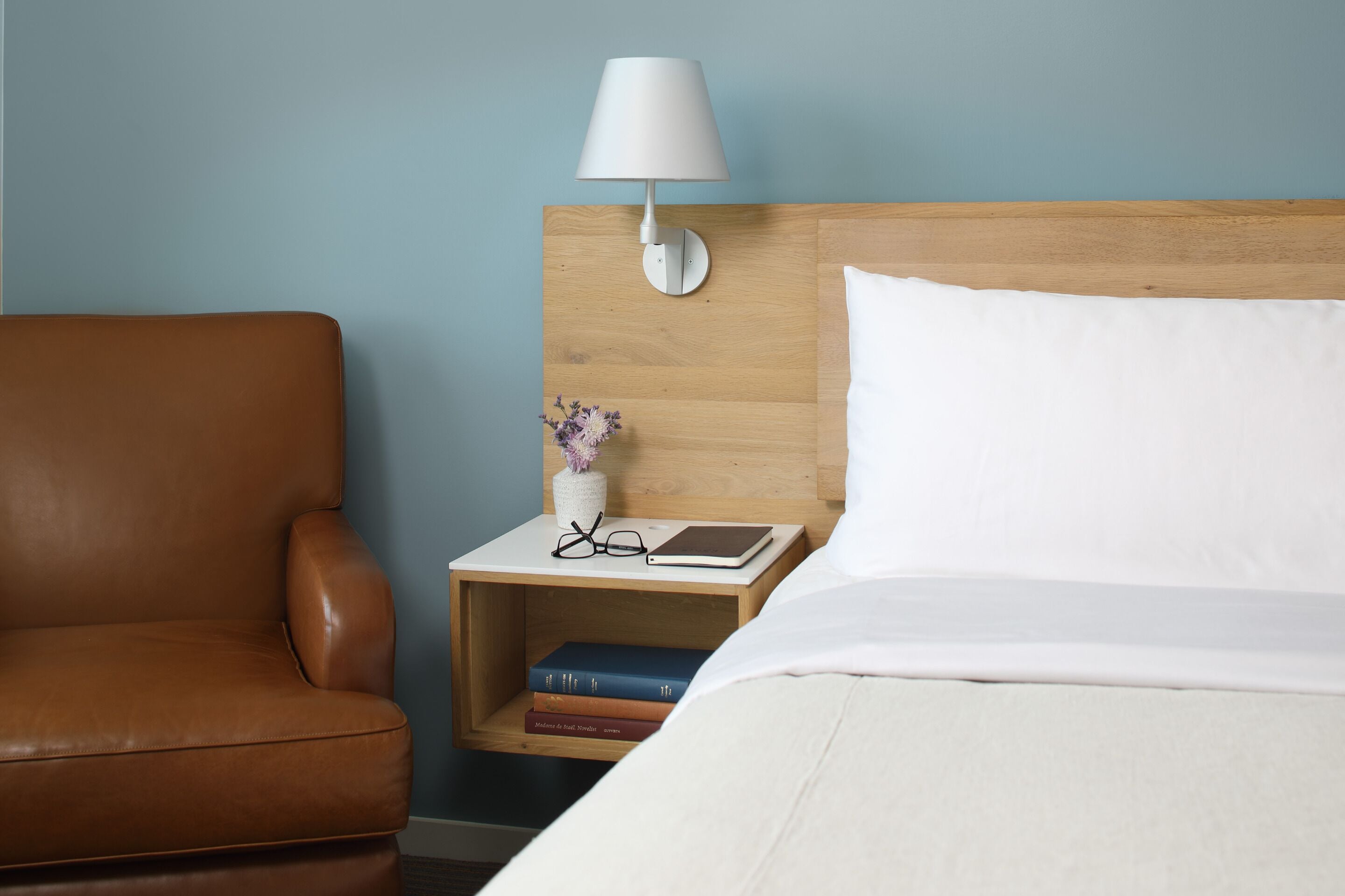 A hotel room with a wooden headboard, a white bed, a brown leather chair, a nightstand with a lamp, and a small vase with flowers.