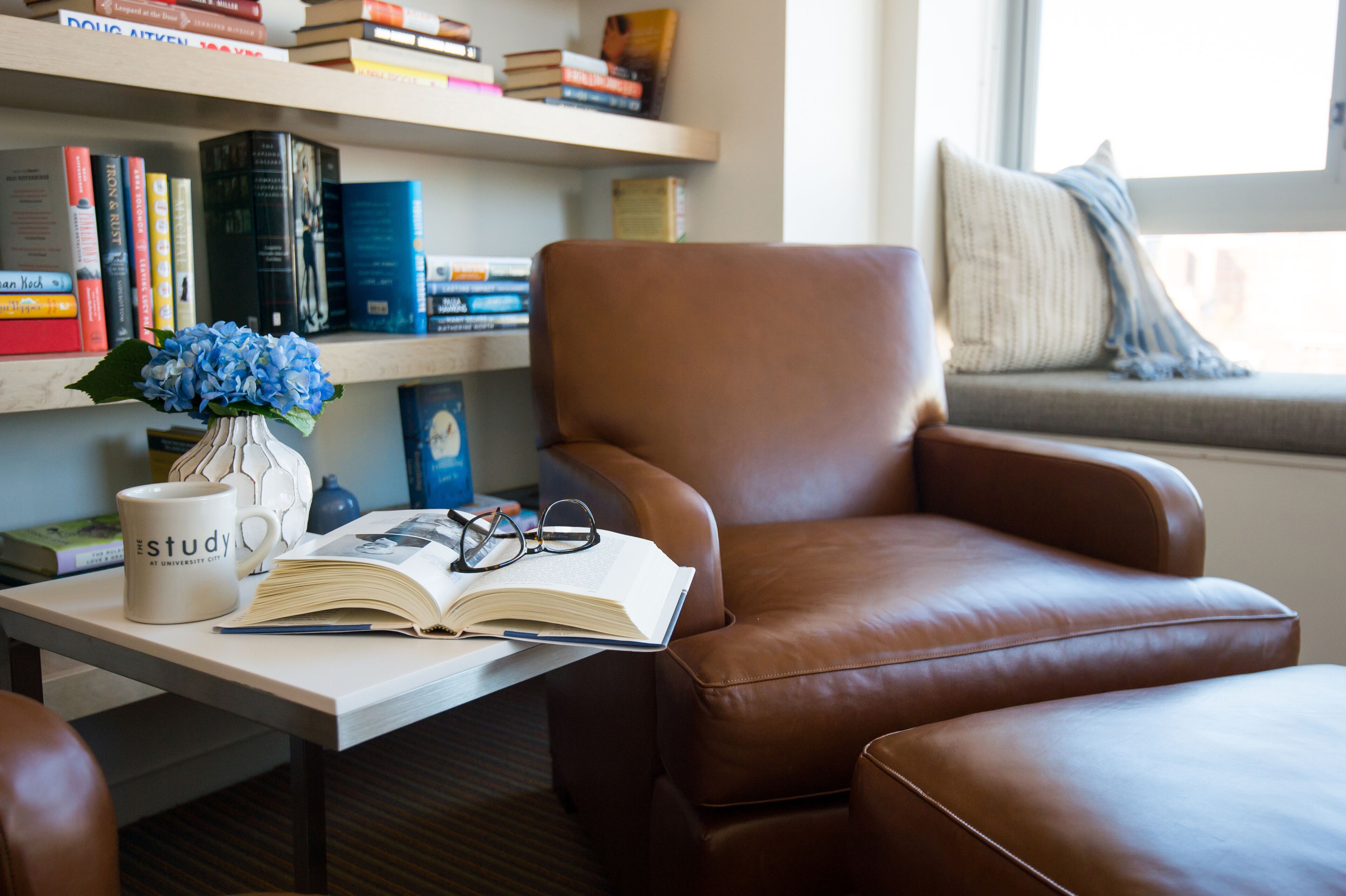 A study area with a brown leather chair, a white mug labeled "study," an open book, and a vase with blue flowers on a table.