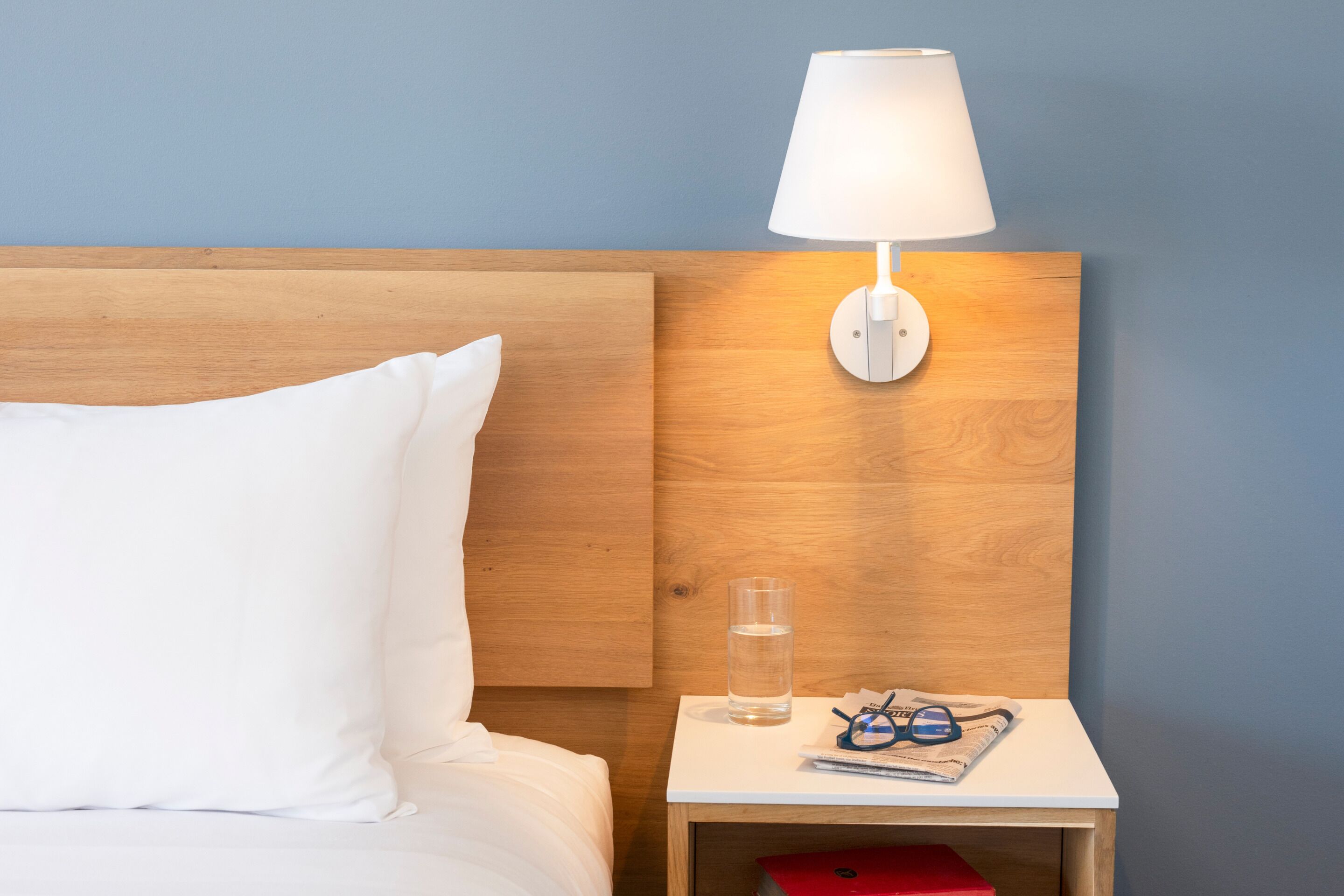 A neatly made bed with white bedding, a wooden headboard, a bedside table with a lamp, a glass of water, and a pair of sunglasses.