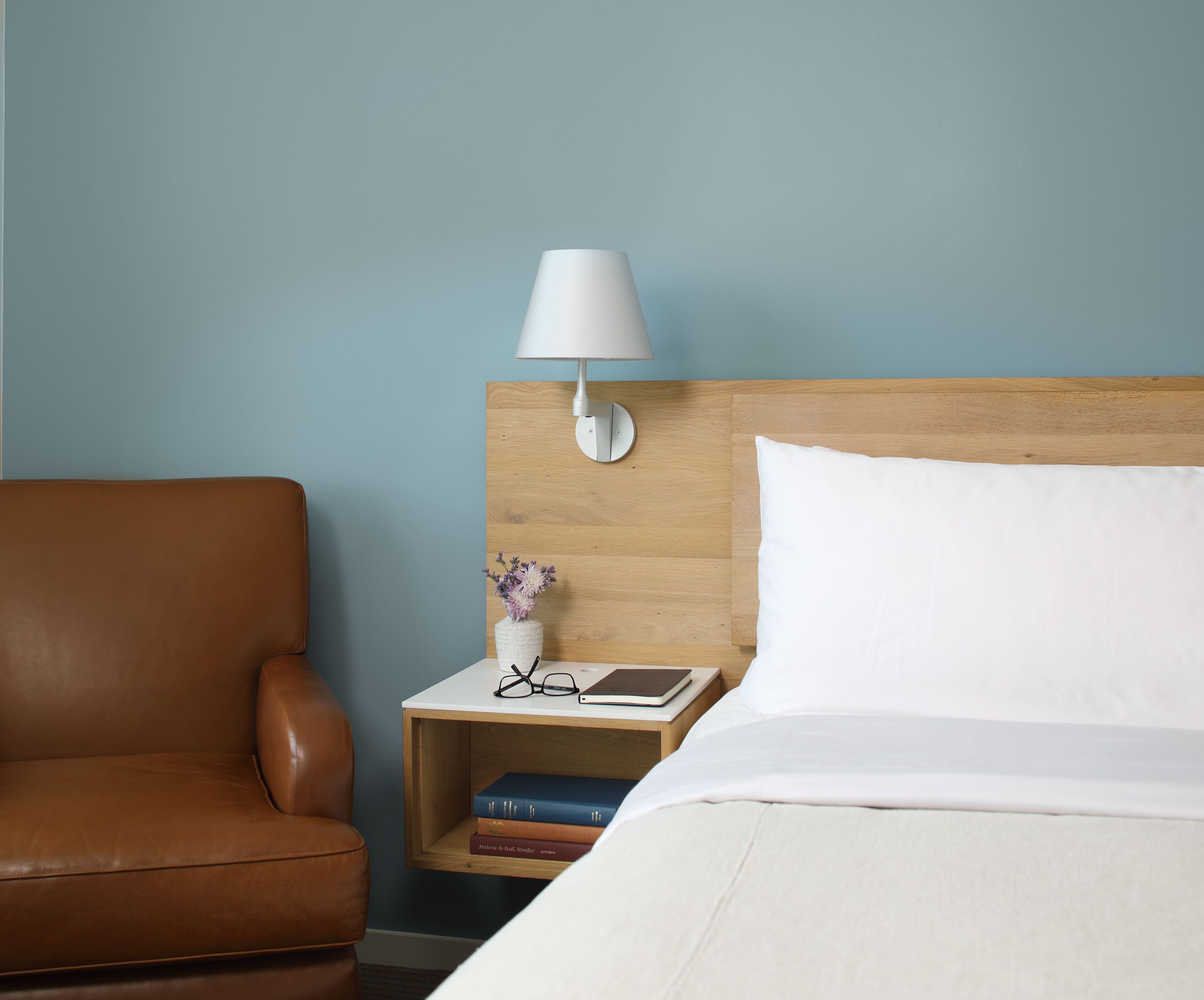 Una habitación de hotel con una butaca de cuero marrón, una mesita de noche de madera con lámpara, libros y un jarrón de flores.