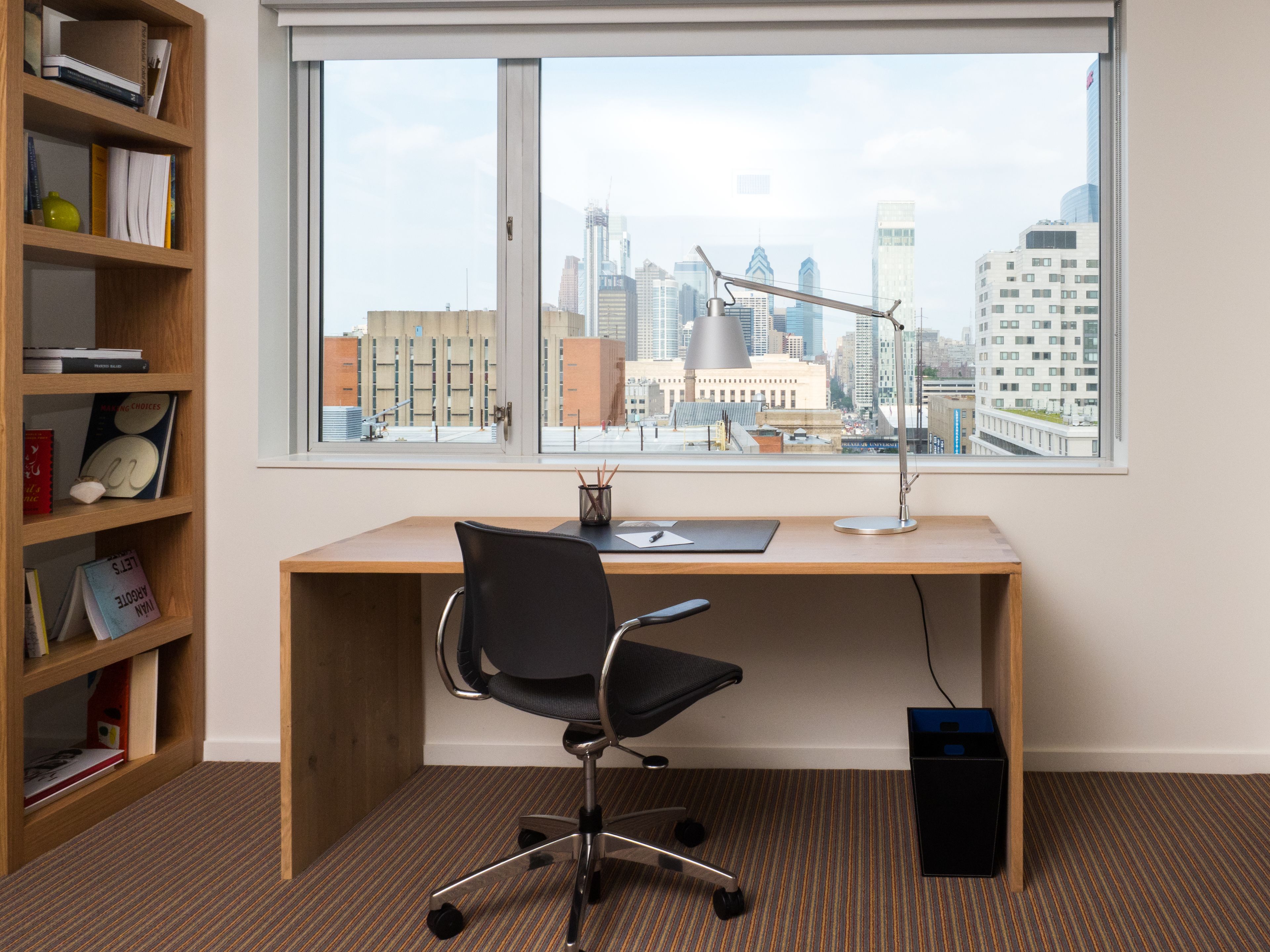 A modern office space with a large window, a desk, a black office chair, and a bookshelf.