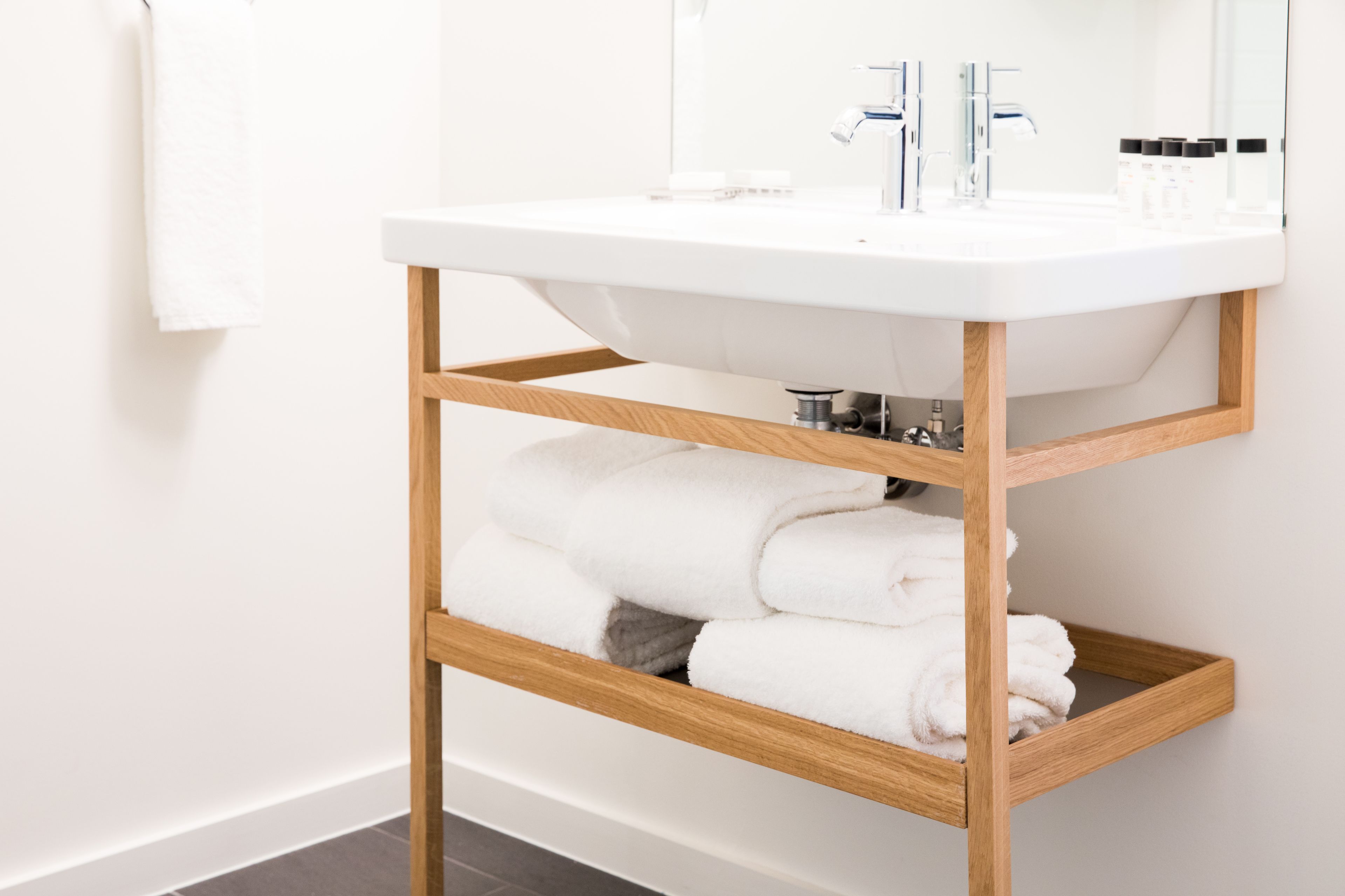 A white bathroom sink with a wooden shelf below holding rolled towels.