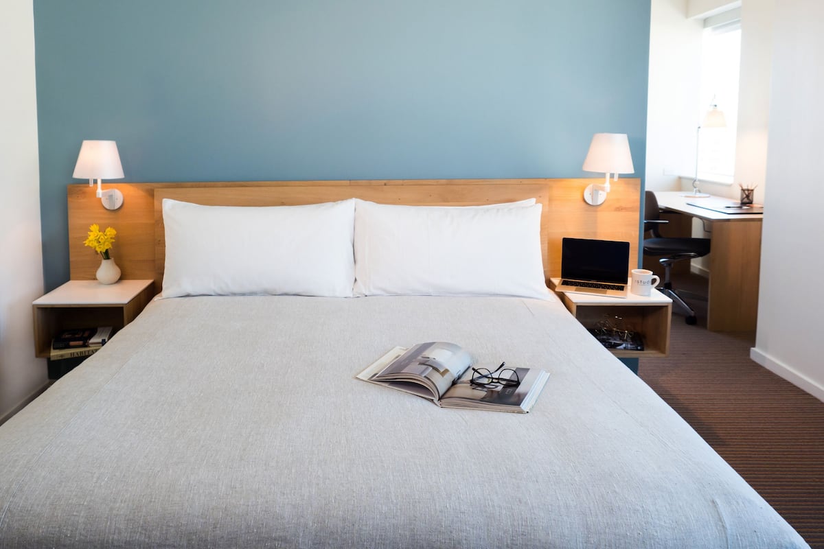 A neatly made bed with white pillows and a magazine, a wooden headboard, two bedside lamps, and a small table with a vase of flowers.