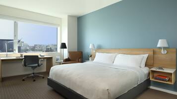 A hotel room with a large bed, a desk with a chair, a laptop, and a view of the city.