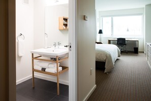 Habitación de hotel con un lavamanos blanco, estantes de madera, una cama, un escritorio y una ventana.