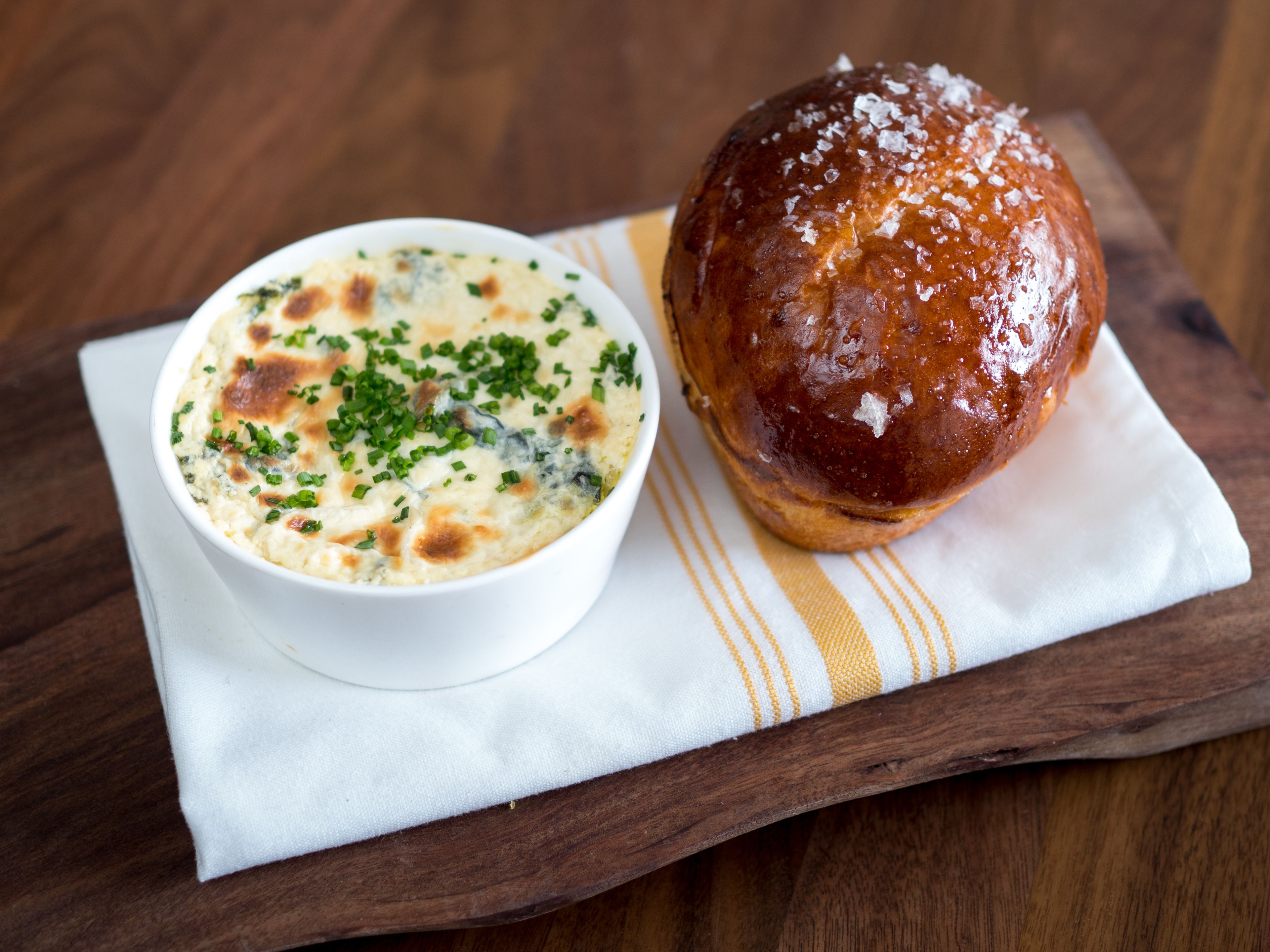 A white bowl of food with a golden-brown top, placed on a wooden table with a striped napkin underneath.