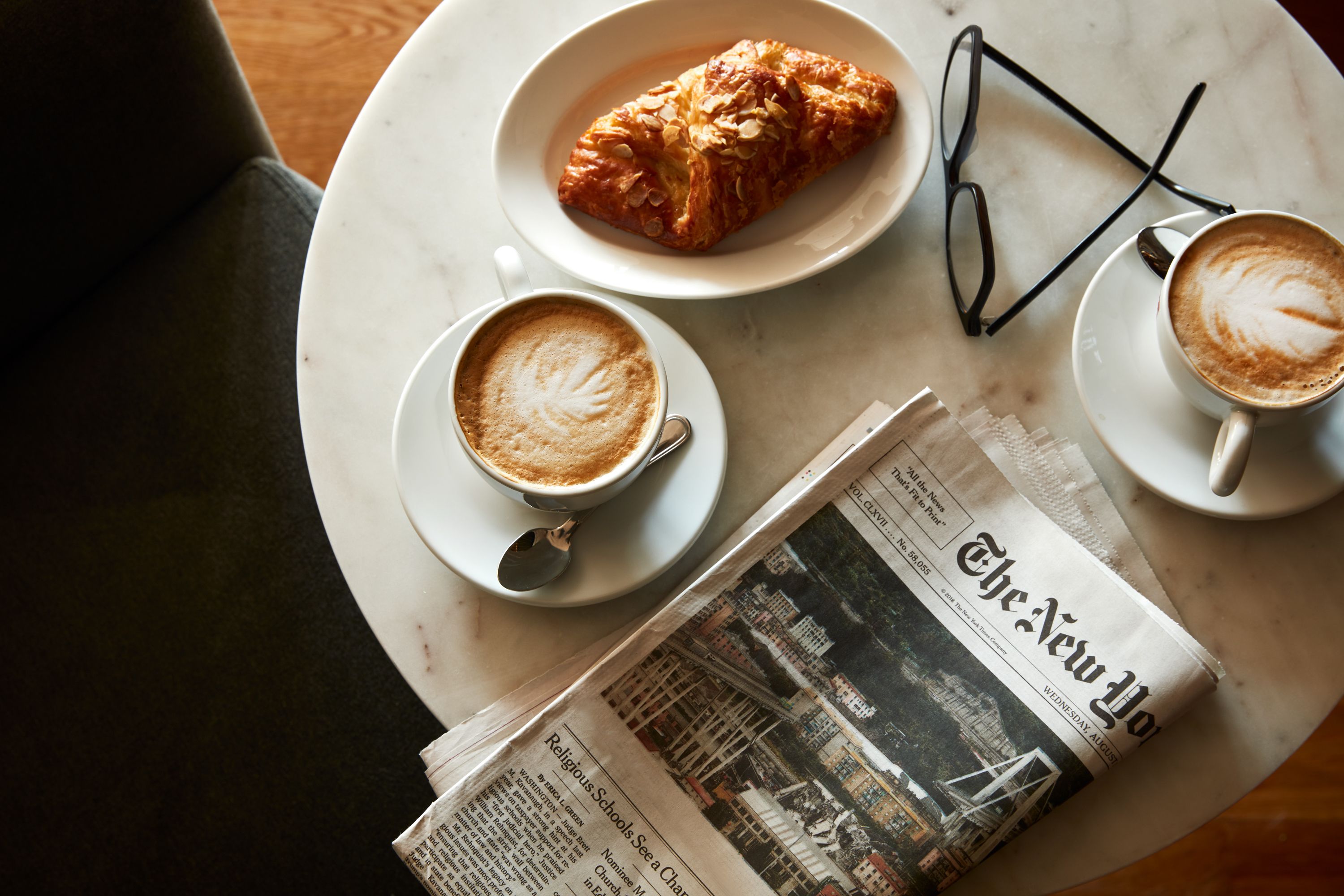 A table with a croissant, two cups of coffee, a pair of glasses, and a newspaper.