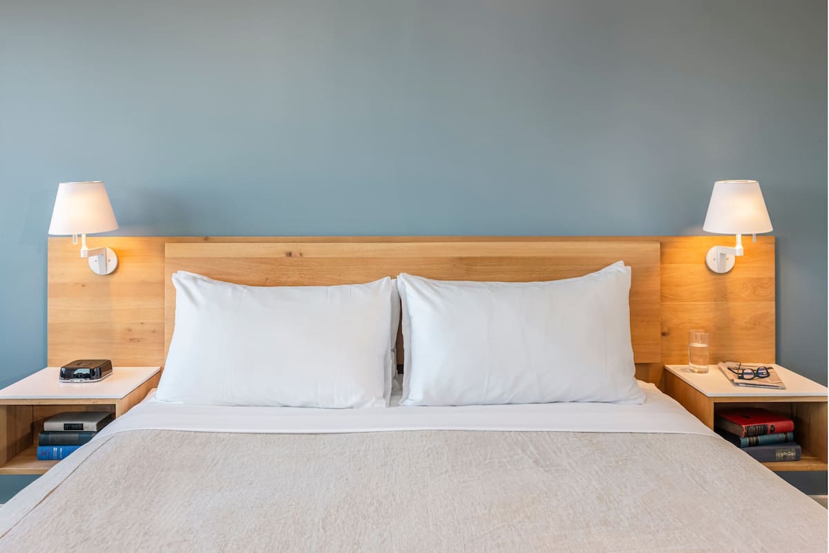A neatly made bed with white linens, two bedside lamps, a wooden headboard, and a nightstand with books and a glass.