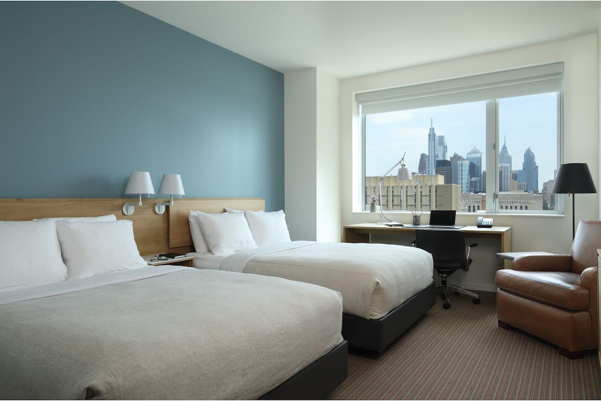 A hotel room with two beds, a desk, a chair, and a large window with a city view.