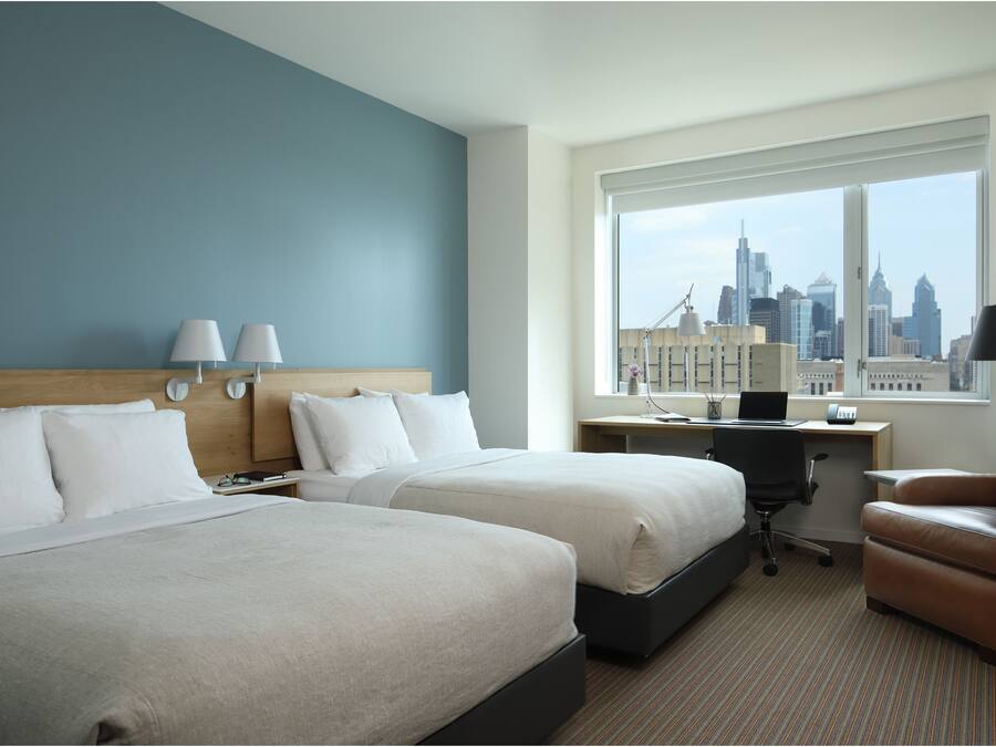 Habitación de hotel con dos camas, un escritorio, una silla y un ventanal con vista a la ciudad.