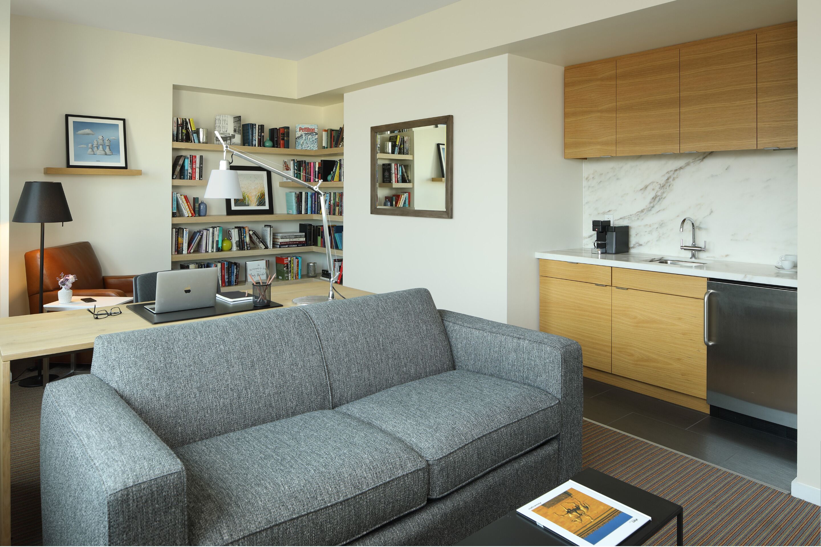 Un salon moderne avec un canapé gris, un bureau avec un ordinateur portable, une bibliothèque et un coin cuisine doté de meubles en bois et d’un plan de travail en marbre.