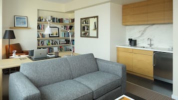Un salon moderne avec un canapé gris, un bureau avec un ordinateur portable, une bibliothèque et un coin cuisine doté de meubles en bois et d’un plan de travail en marbre.