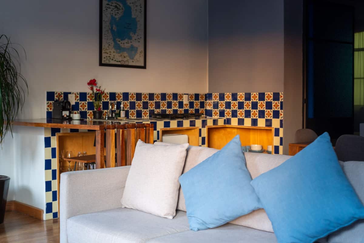 A living room with a sofa, blue cushions, a kitchen area with a tiled backsplash, and a dining table.