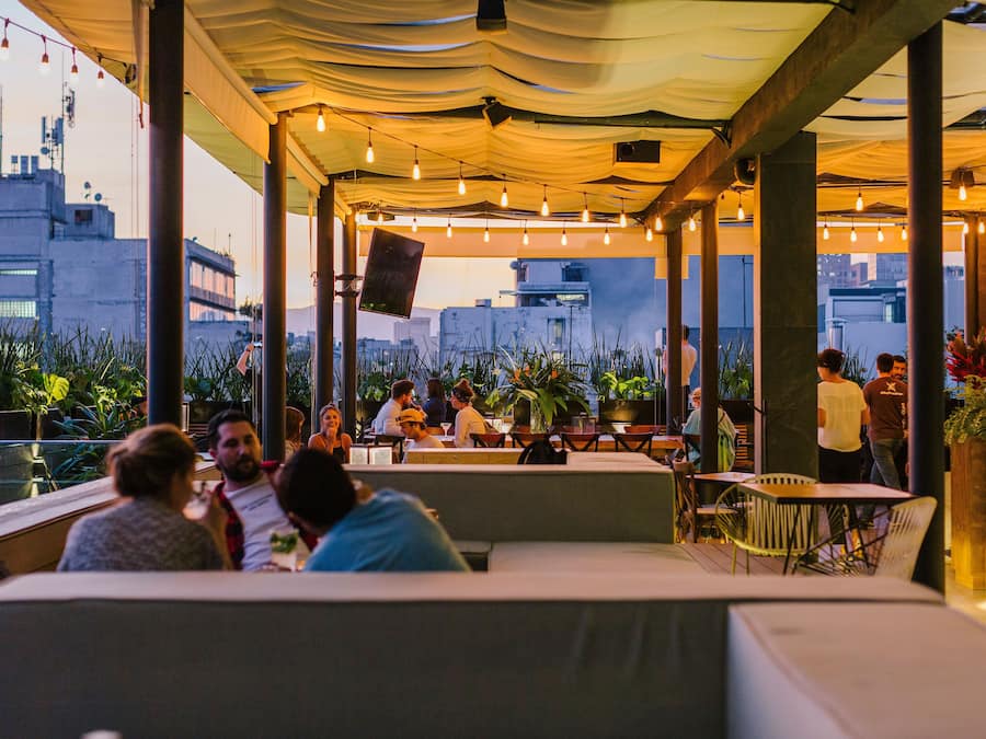 An outdoor seating area with tables and chairs, surrounded by potted plants and a view of buildings.
