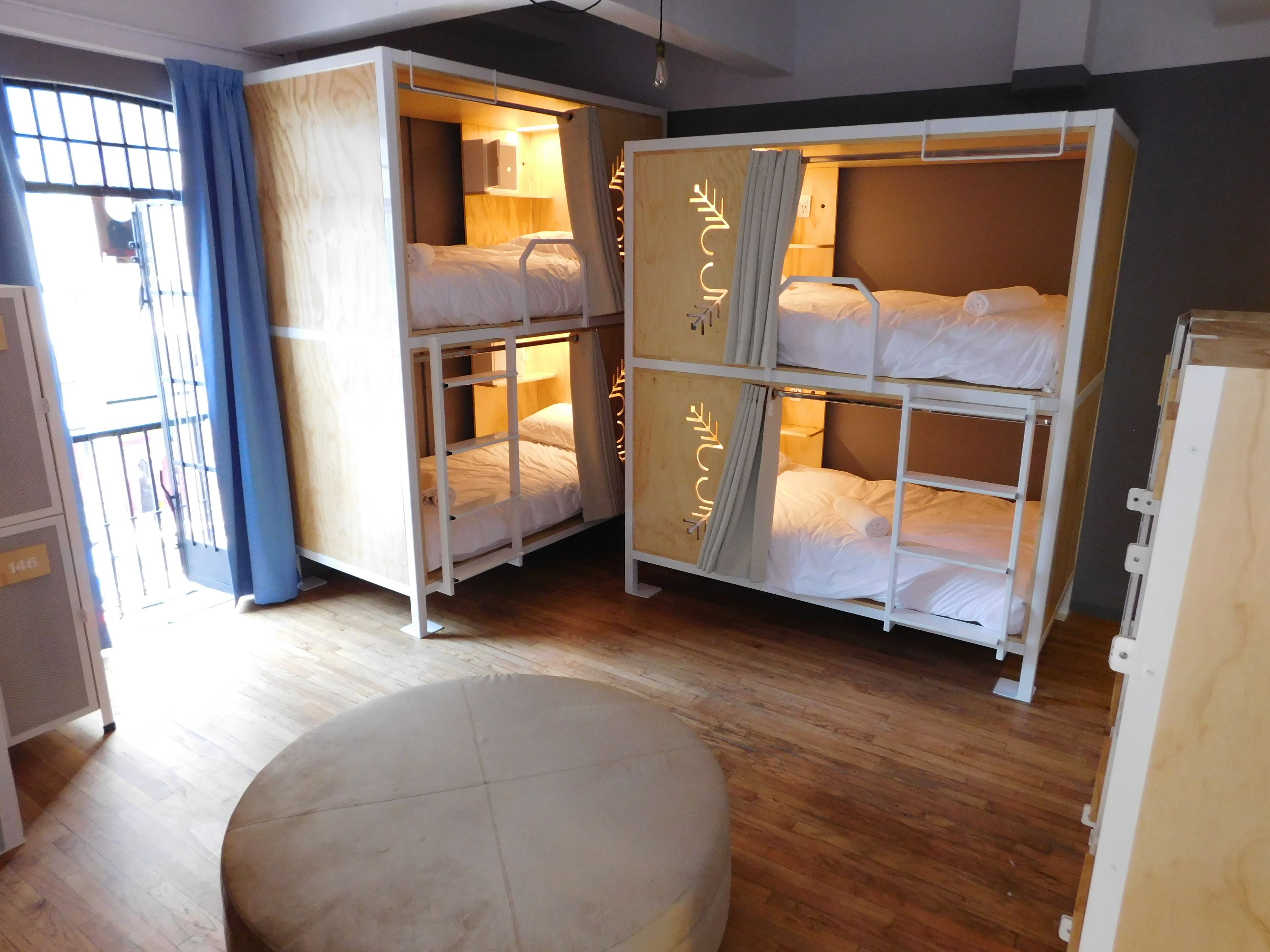 A dormitory room with bunk beds, a wooden floor, and a window with blue curtains.
