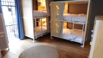 A dormitory room with bunk beds, a wooden floor, and a window with blue curtains.