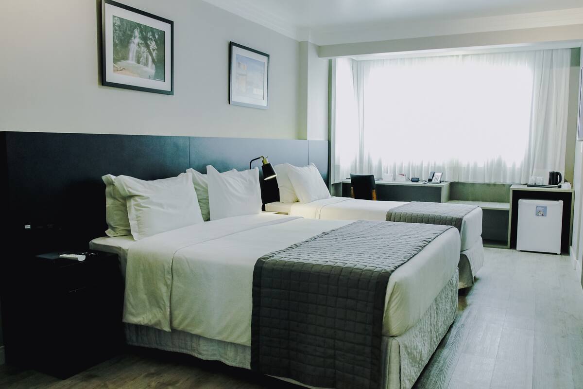 A hotel room with two beds, a desk, a small fridge, and a window with curtains.