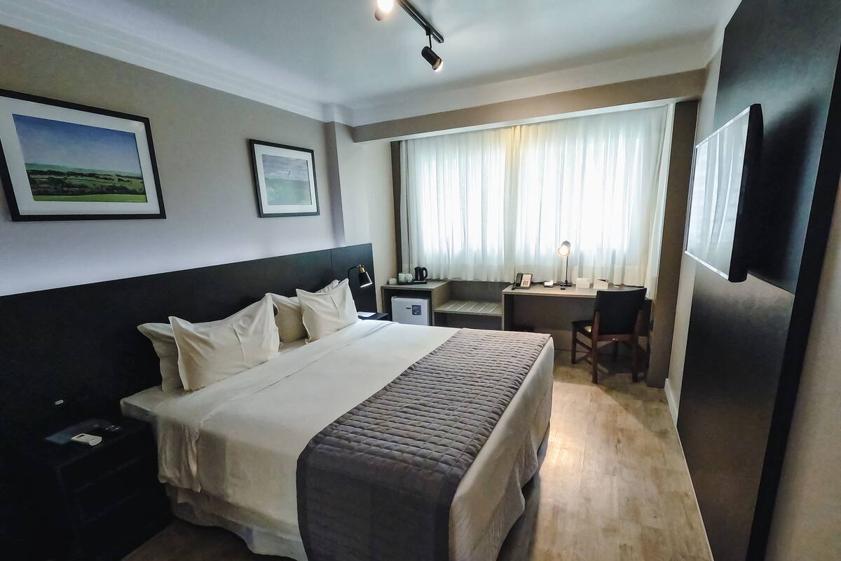 A hotel room with a large bed, a desk, a chair, a small refrigerator, and two framed pictures on the wall.