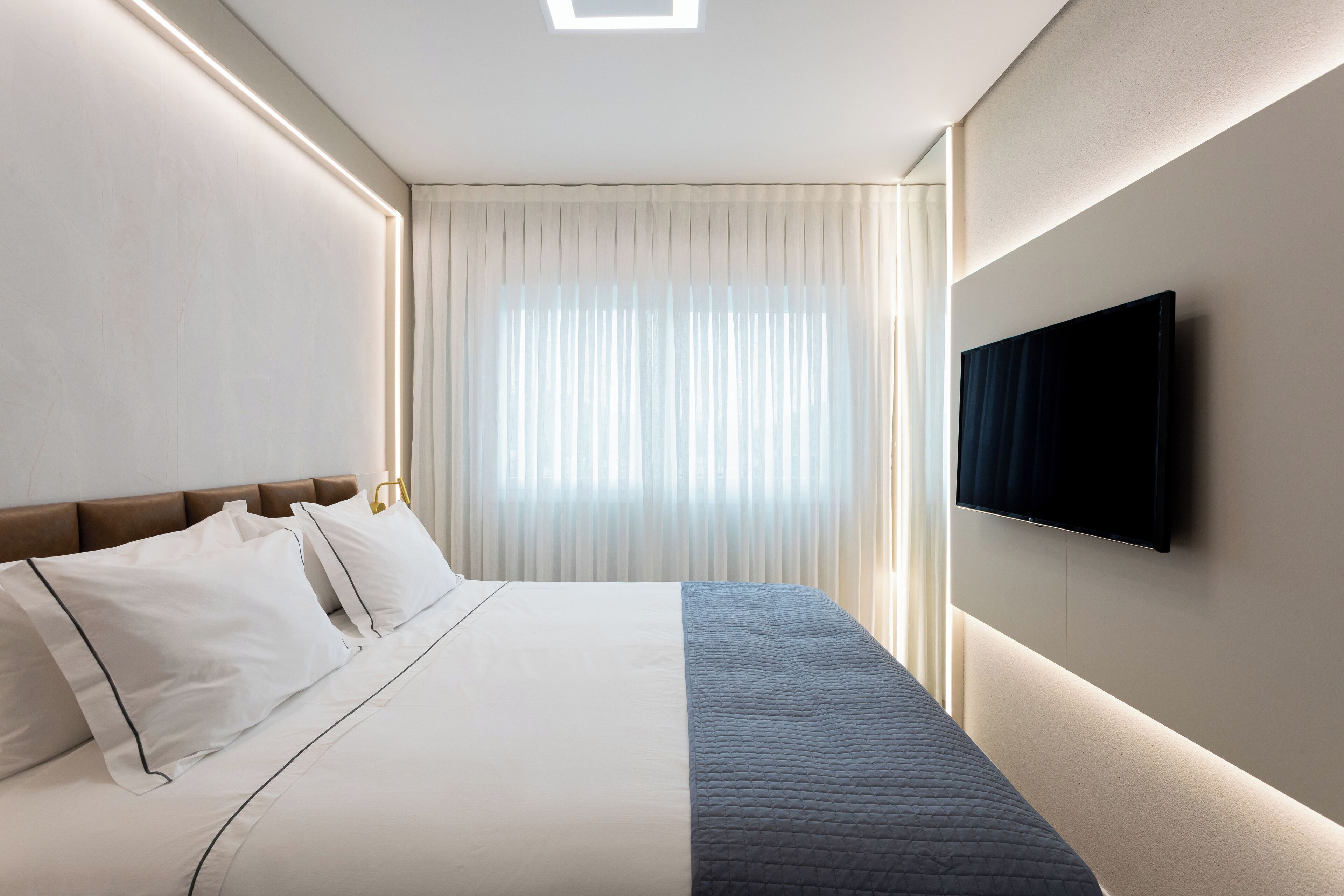Una habitació d'hotel moderna amb un llit ben fet, un televisor de pantalla plana fixat a la paret i un gran finestral amb cortines transparents.