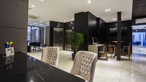 A modern interior with a marble reception desk, cushioned chairs, a potted plant, and a visible elevator. - Santa Inn Hotel (Balneário Camboriú)