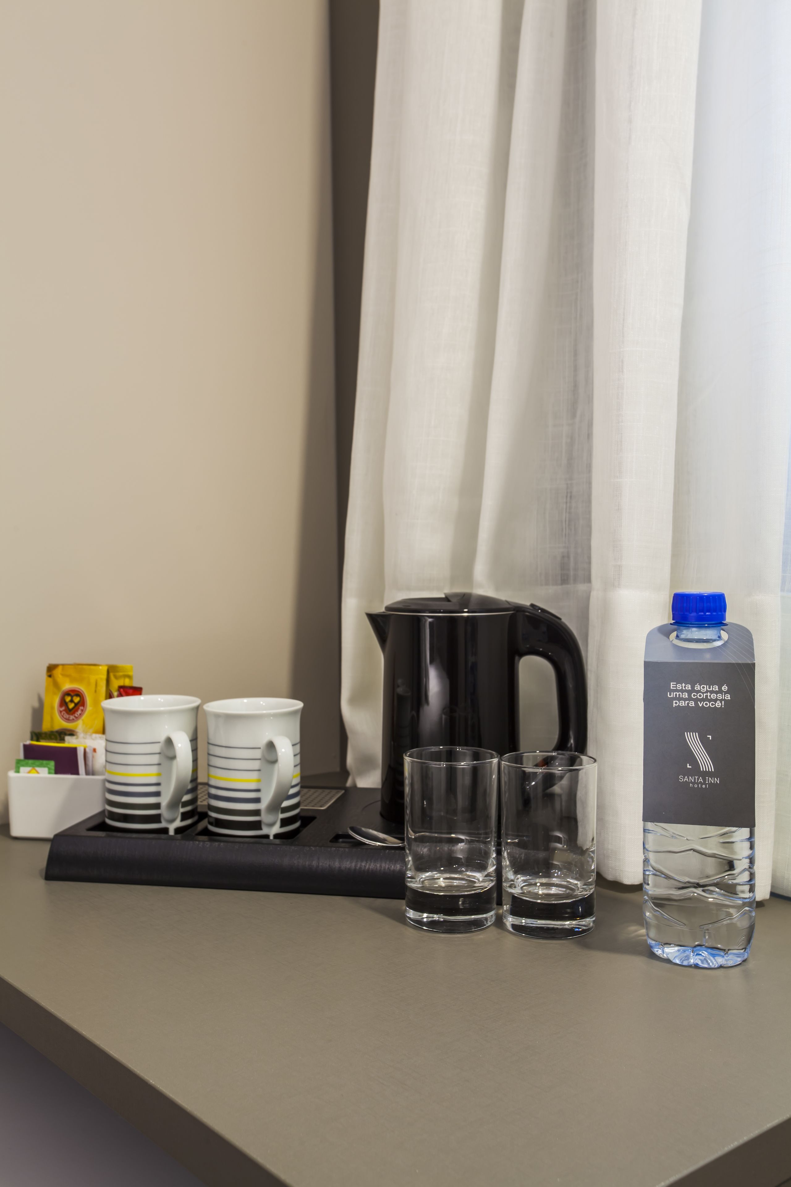 Ontbijt in een hotelkamer, met een dienblad waarop twee witte mokken met strepen, twee glazen, een zwart elektrisch waterkoker, een waterfles en een pakje koffiemelk staan.