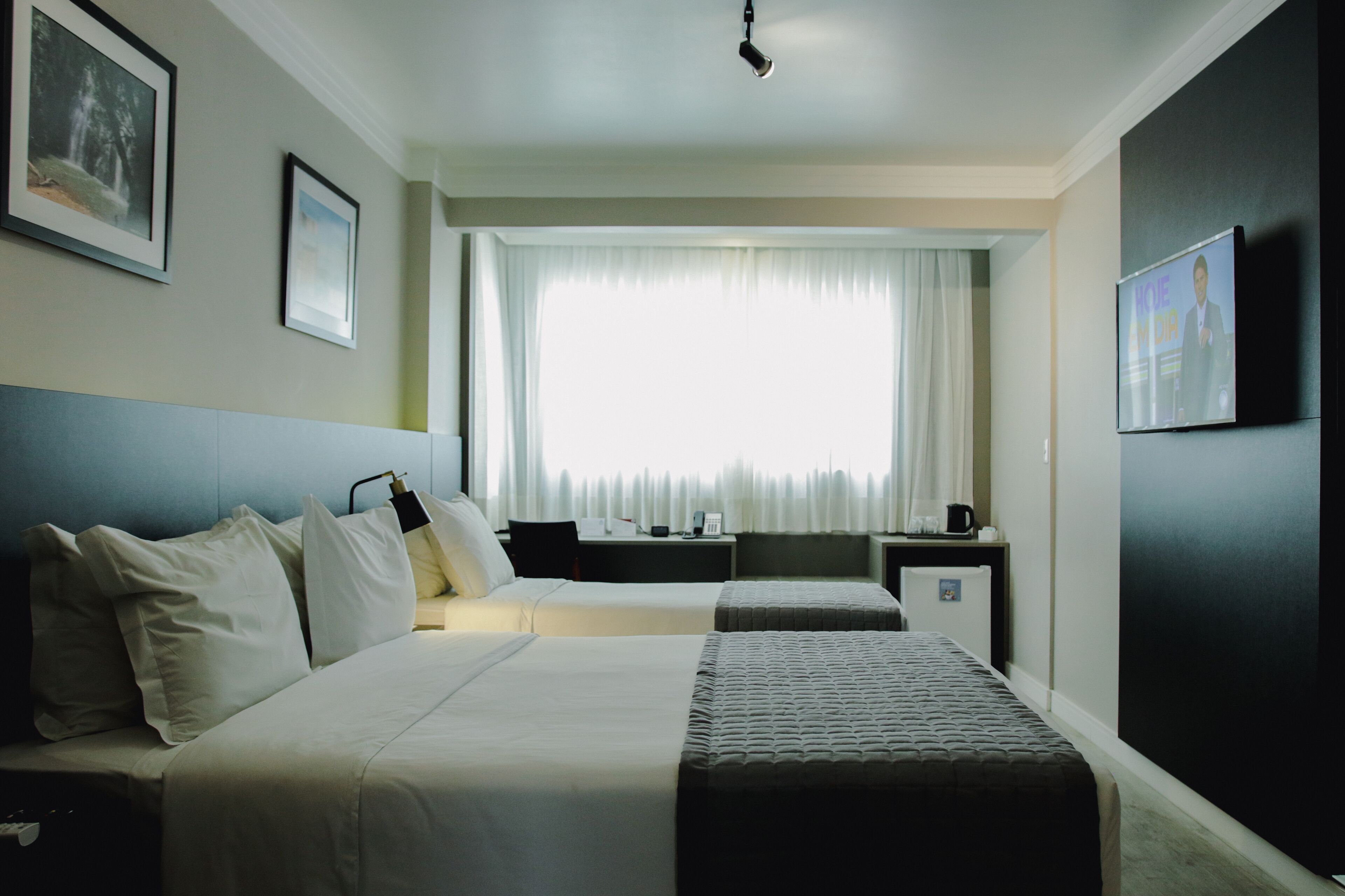A hotel room with two beds, a desk, a chair, a TV, and a window with curtains.