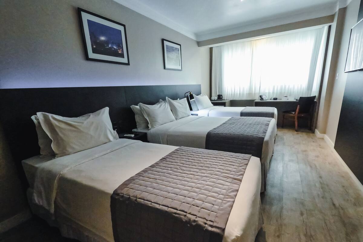 A hotel room with two beds, a desk, and a chair. There are framed pictures on the wall and a window with curtains.