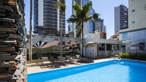 A swimming pool with lounge chairs and umbrellas, surrounded by tall buildings and palm trees. - Santa Inn Hotel (Balneário Camboriú)