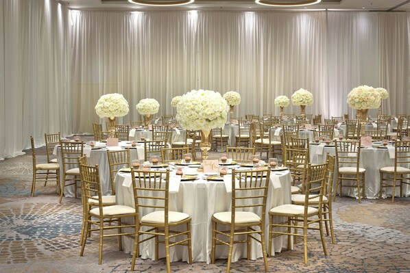 Un salón de banquetes con mesas redondas, sillas doradas y centros de mesa florales blancos.