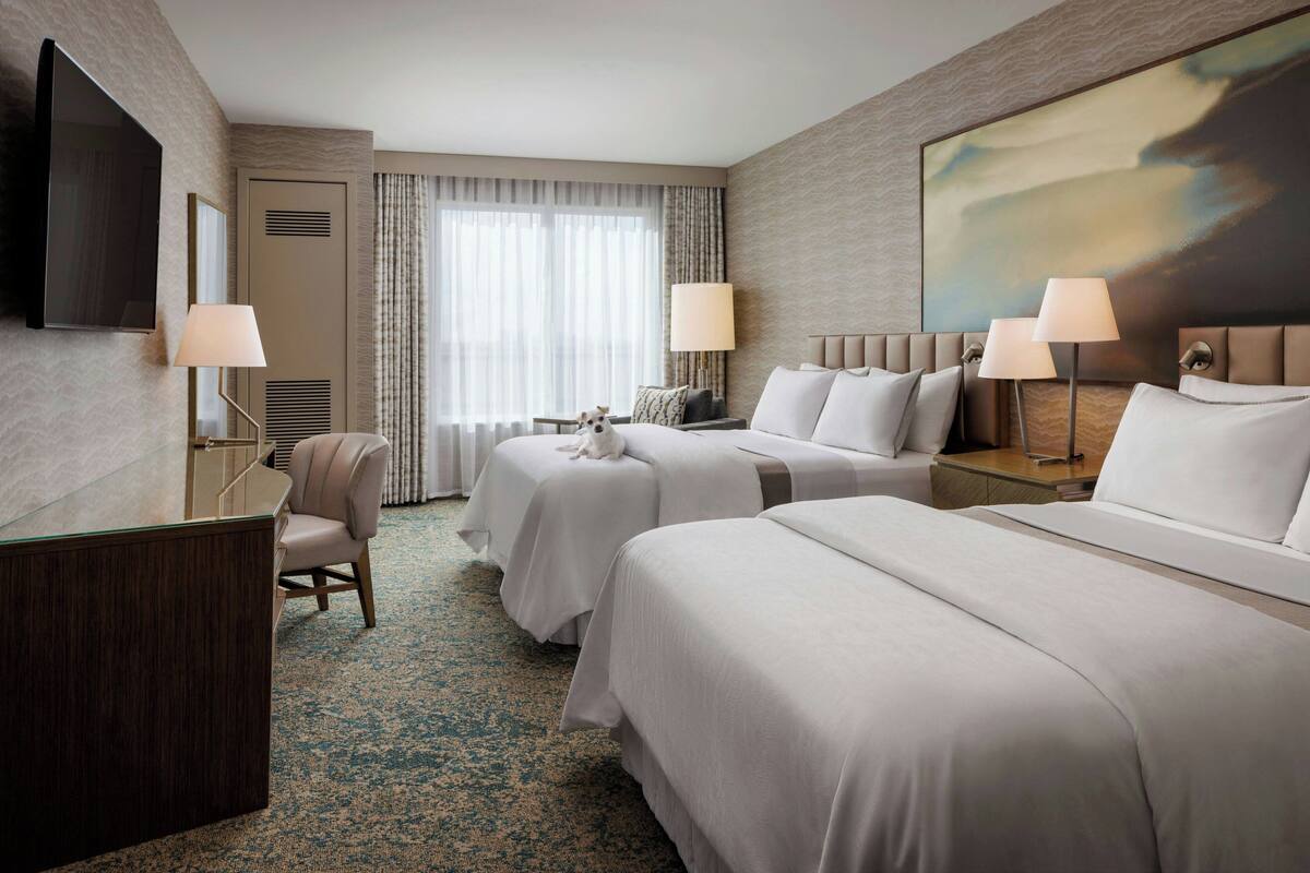 A hotel room with two beds, a desk, a chair, a TV, and a large painting on the wall.