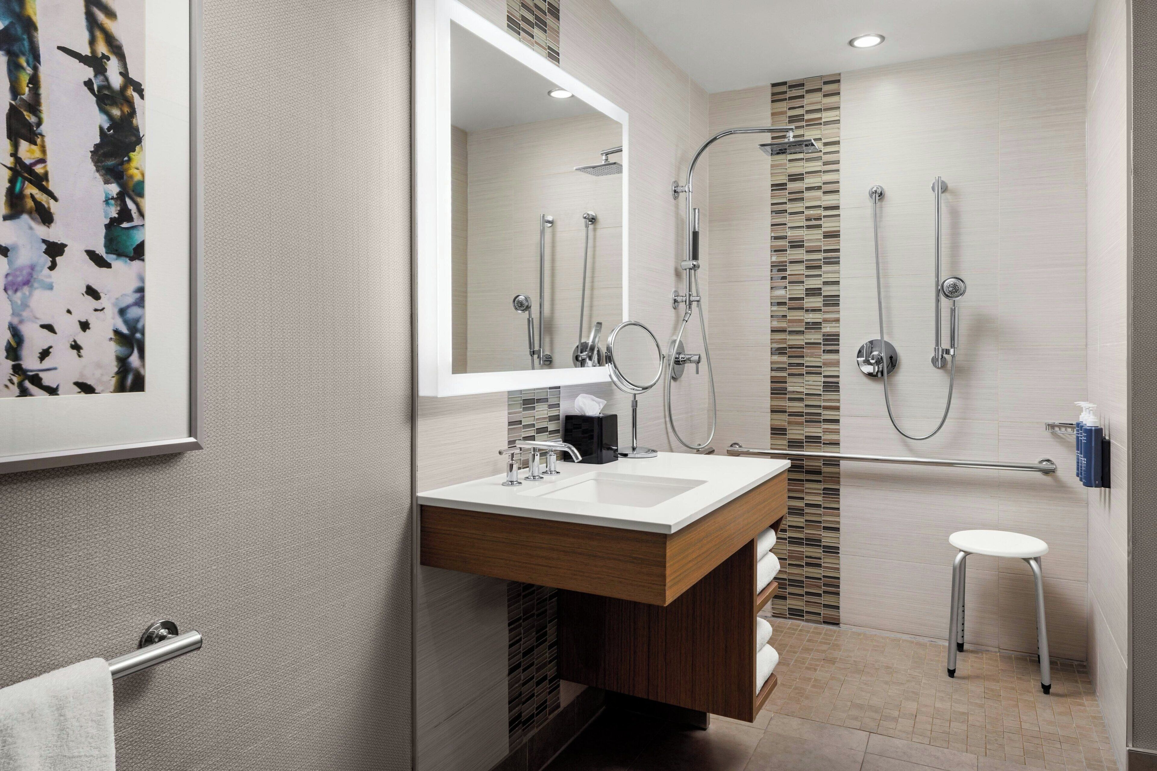 A modern bathroom with a large mirror, a sink, a shower with a glass door, and a small stool.