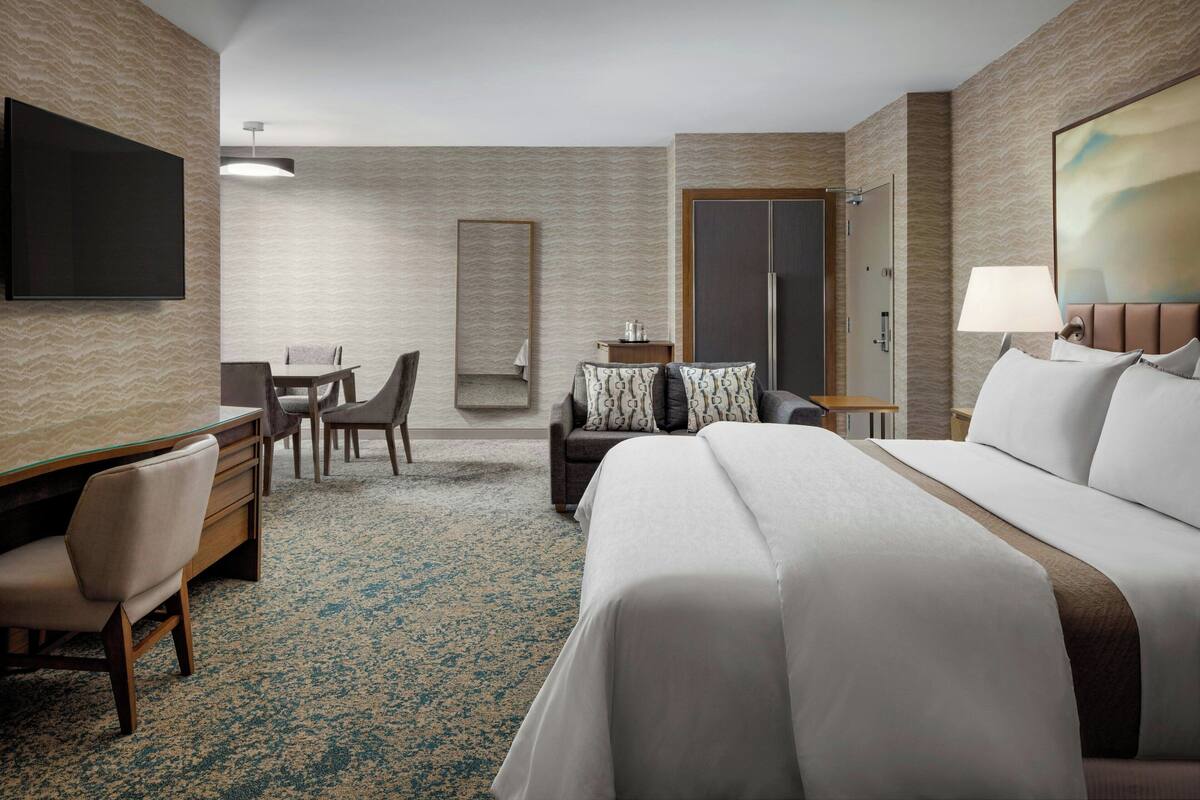 A hotel room with a large bed, a desk with a chair, a small table with a chair, a TV mounted on the wall, and a door.