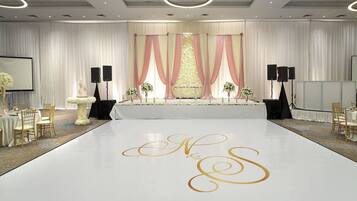 A wedding reception area with a white dance floor, a stage with draped curtains, and a floral arrangement.