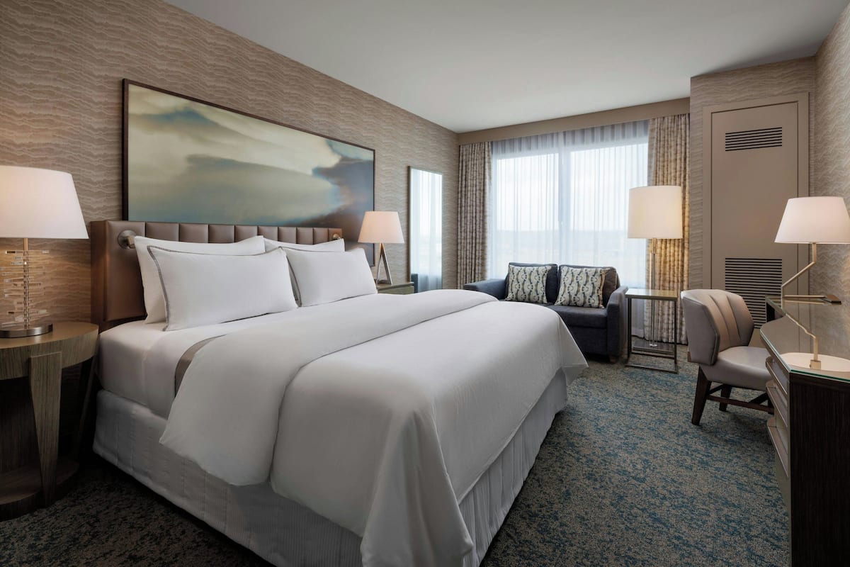 A hotel room with a large bed, two bedside tables, a desk, a chair, a sofa, and a large painting on the wall.
