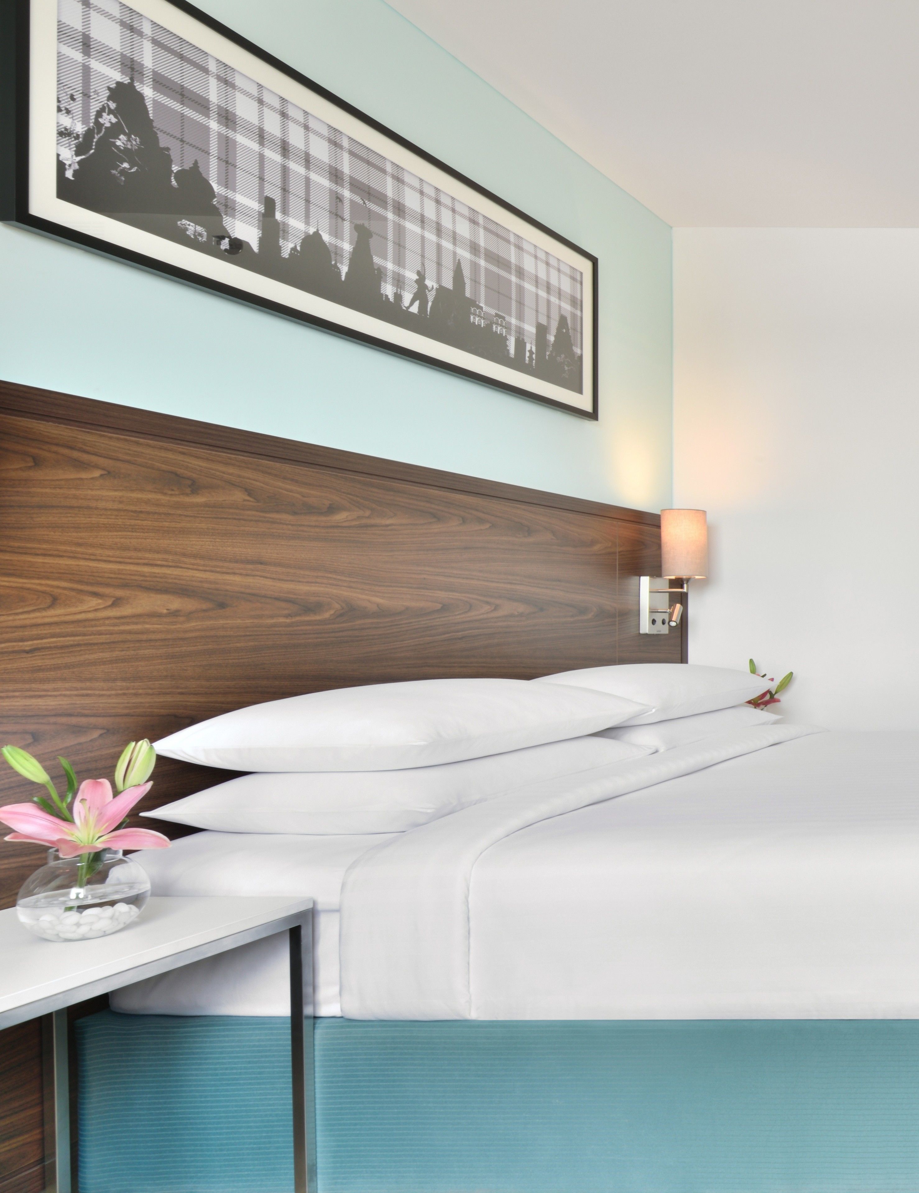 Una cama bien hecha con sábanas blancas, cabecera de madera, un cuadro enmarcado encima, una lámpara de noche y un florero con flores sobre una mesita de noche.
