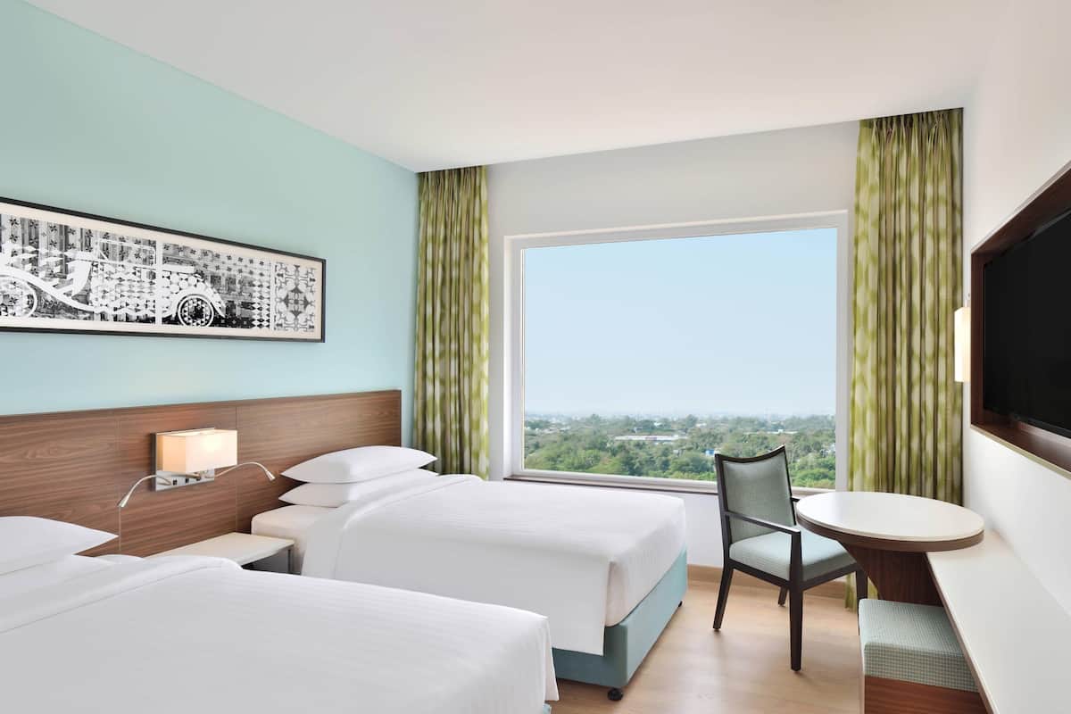 Une chambre d’hôtel avec deux lits, un bureau, une chaise, une télévision et une grande fenêtre donnant sur la ville.