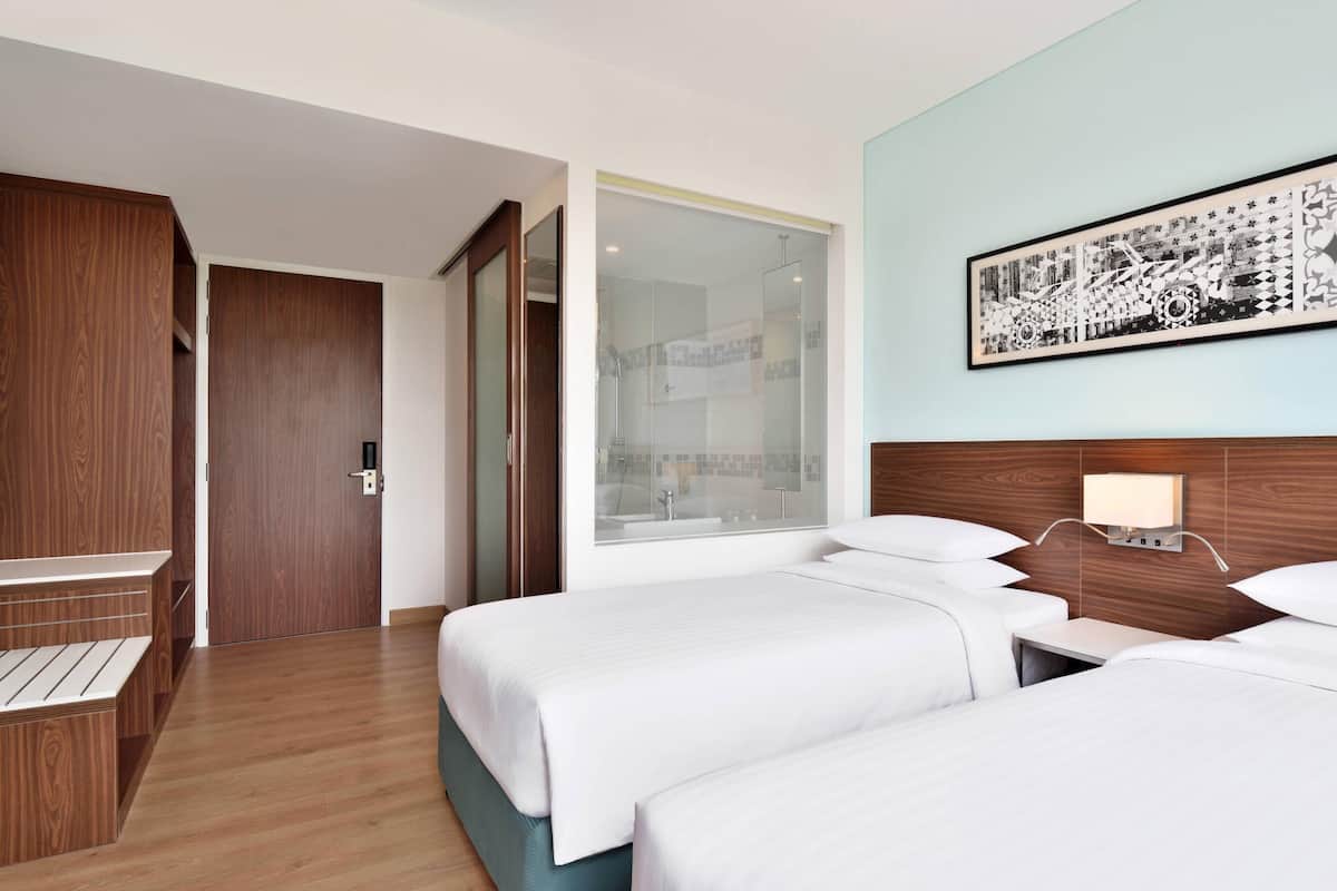 Une chambre d’hôtel avec deux lits, une armoire en bois, une cabine de douche en verre et un luminaire fixé au mur.
