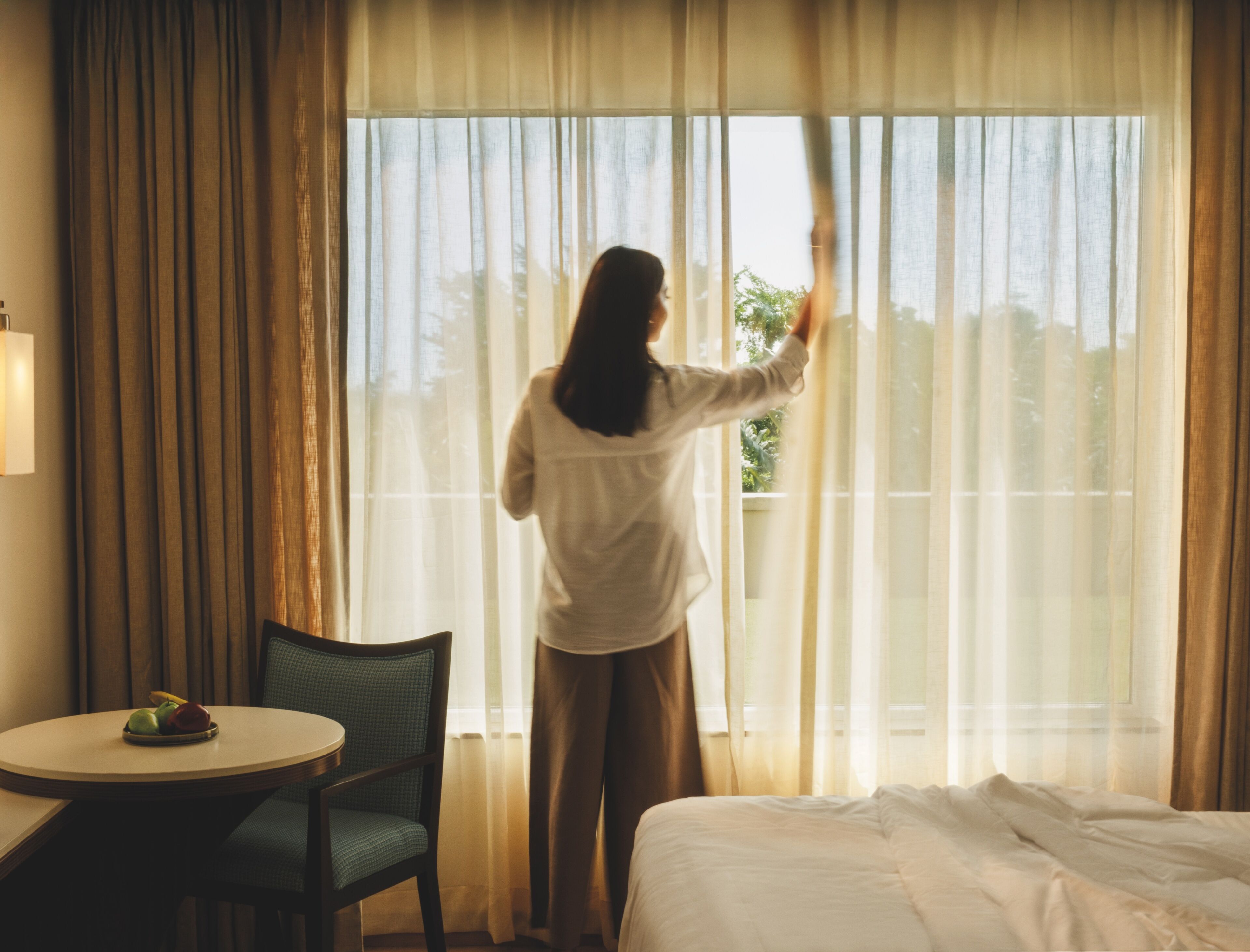 Une chambre d’hôtel avec un lit, une chaise, une petite table avec des fruits et une fenêtre avec des rideaux.