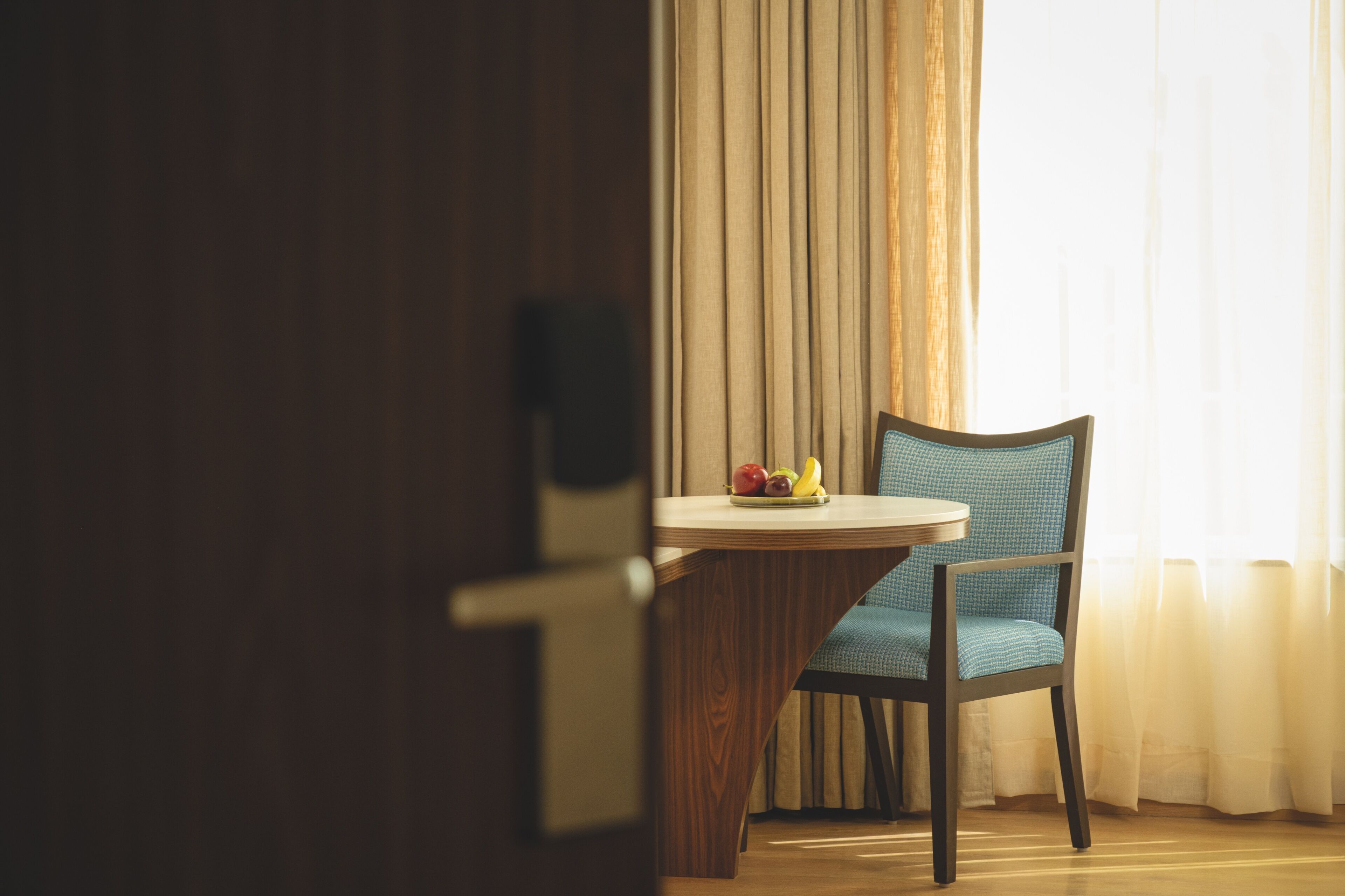 Una habitación con puerta de madera, una mesa redonda con frutero, una silla azul y una ventana con cortinas.