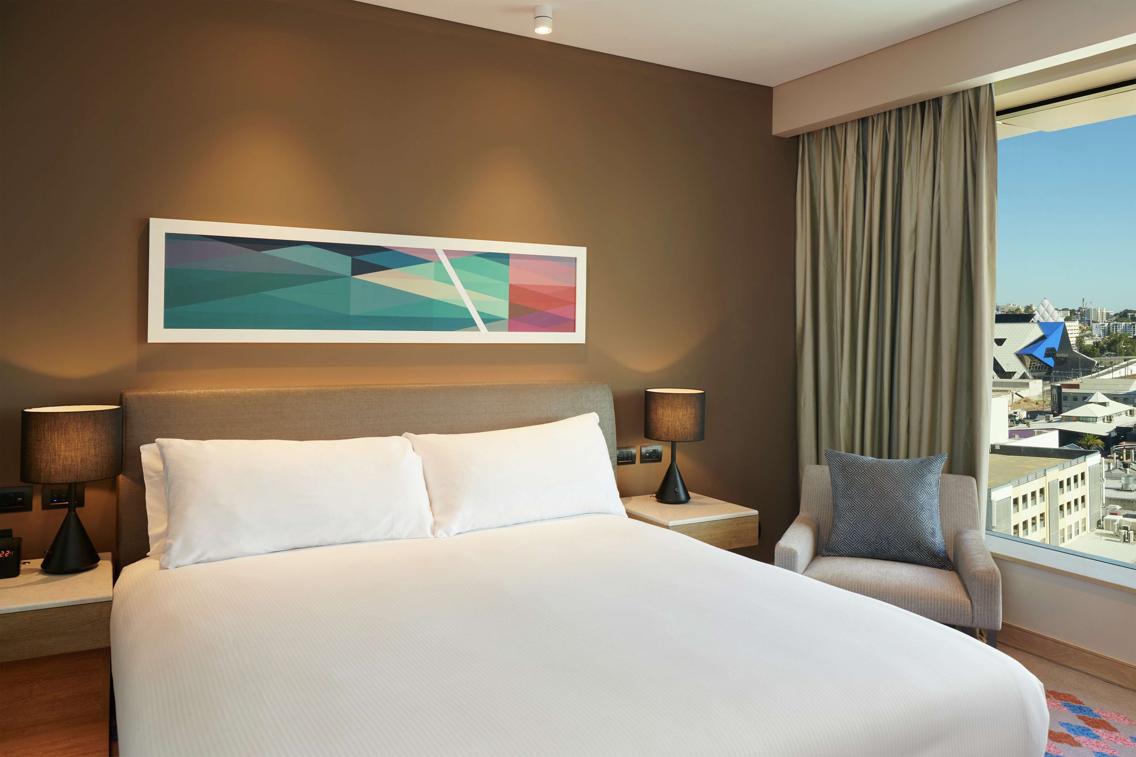 A hotel room with a large bed, two bedside tables with lamps, a chair with a pillow, a large window with a city view, and a colorful abstract painting on the wall.