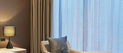 A hotel room with a large bed, a chair, a small table, and a window with curtains.