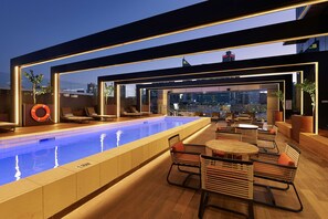 Un área de piscina en la azotea con vistas al horizonte urbano, sillones de descanso y mesas.