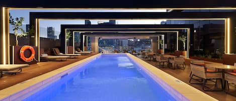 An indoor swimming pool with lounge chairs and tables, surrounded by glass walls offering city views.