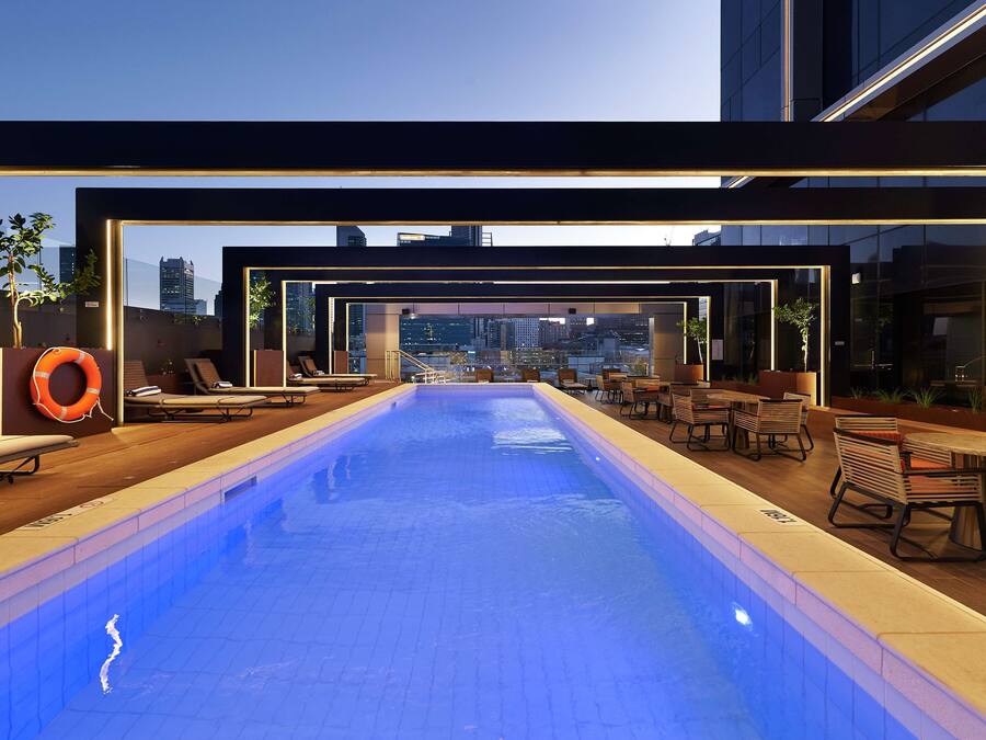 An indoor swimming pool with lounge chairs and tables, surrounded by glass walls offering city views.