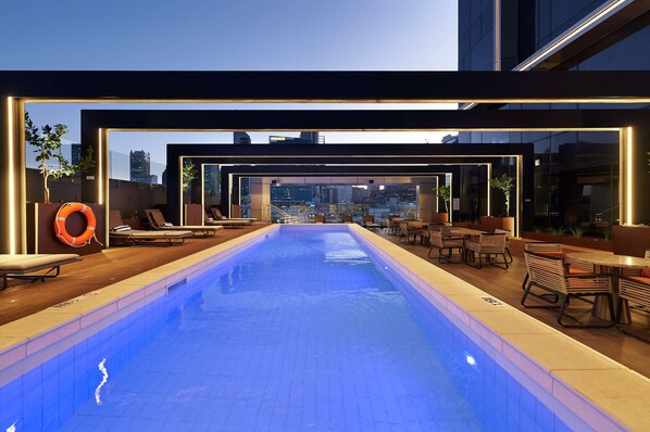 Una piscina interior con sillones y mesas, rodeada de paredes de cristal que ofrecen vistas de la ciudad.