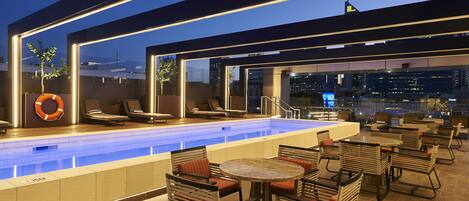 A rooftop pool area with lounge chairs and tables, surrounded by glass walls offering city views.